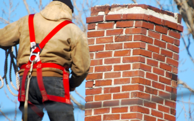 A worker in a harness inspects a brick chimney on a roof against a clear blue sky.