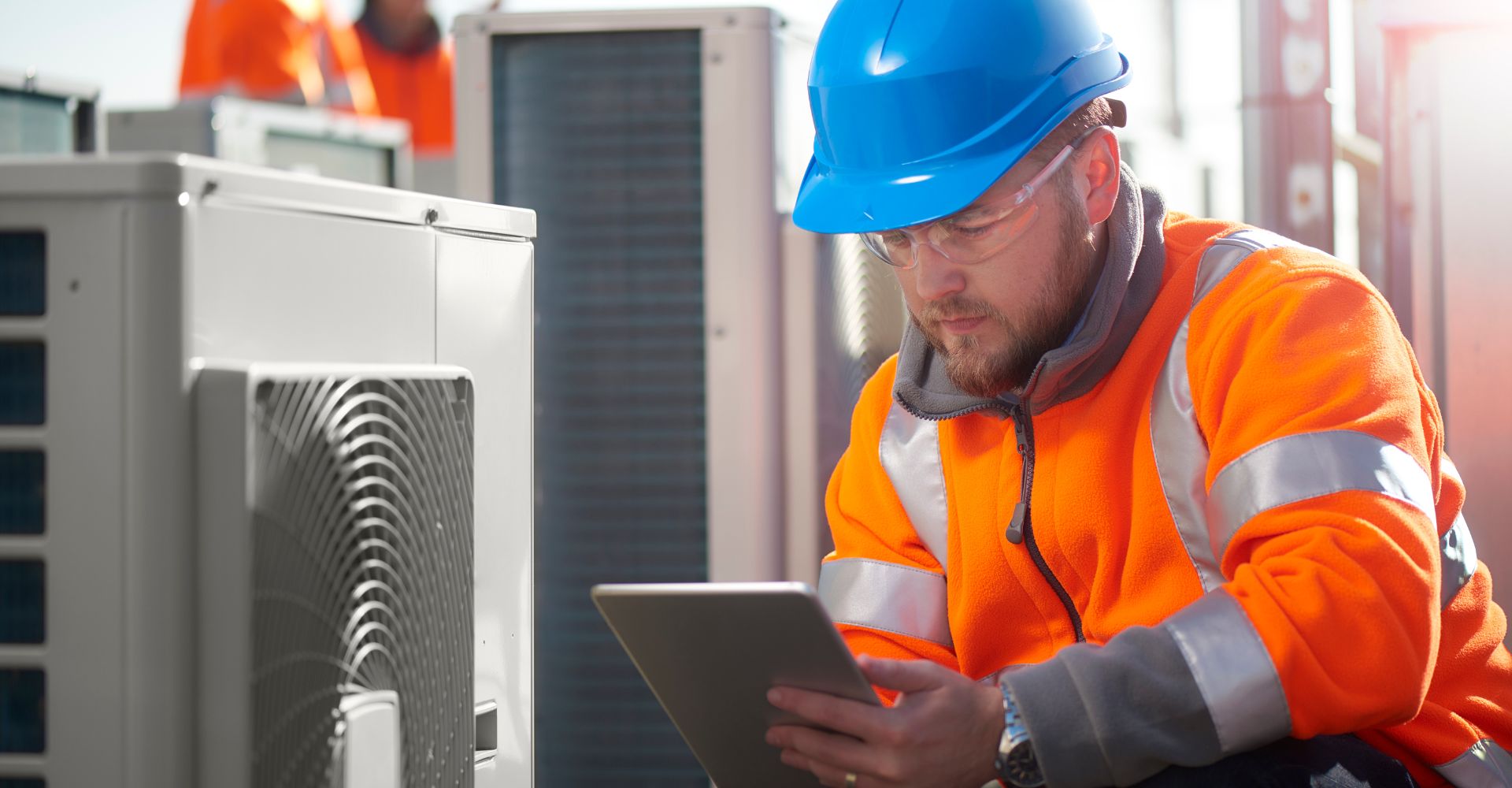 Worker in orange jacket and blue hard hat using a tablet near HVAC units.