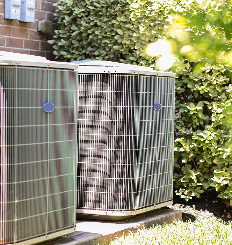 Outdoor HVAC units next to a brick wall, surrounded by greenery.