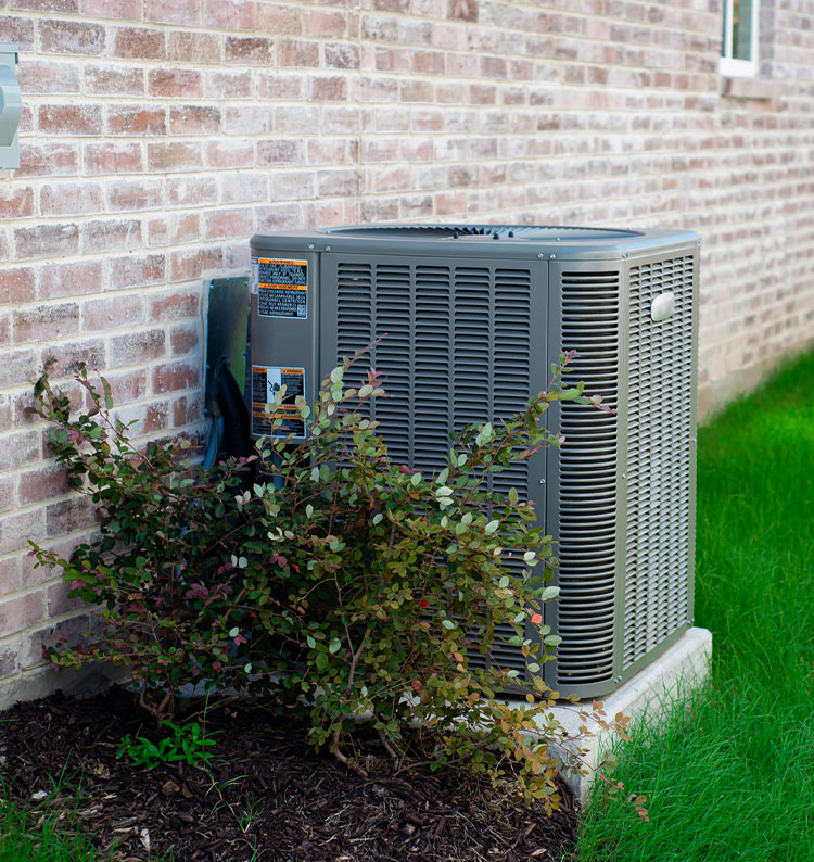Outdoor air conditioning unit beside brick wall with green foliage.