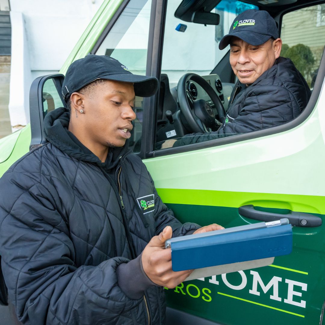 Two men in Clover-branded jackets review a clipboard beside a green van.