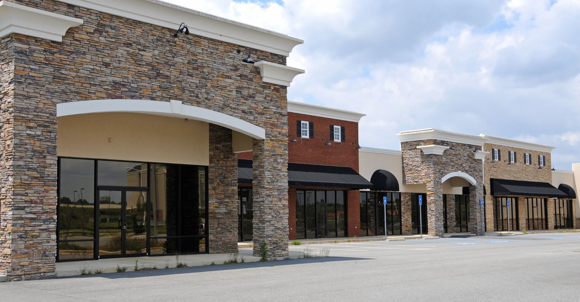 Empty retail strip mall with stone facade and large glass storefronts under a partly cloudy sky.