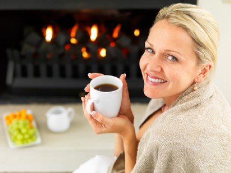 Woman enjoying coffee by the fireplace, wrapped in a blanket, with a plate of fruit nearby.