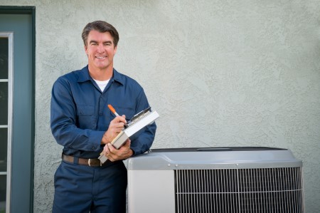 Technician in blue uniform with clipboard inspects outdoor HVAC unit.