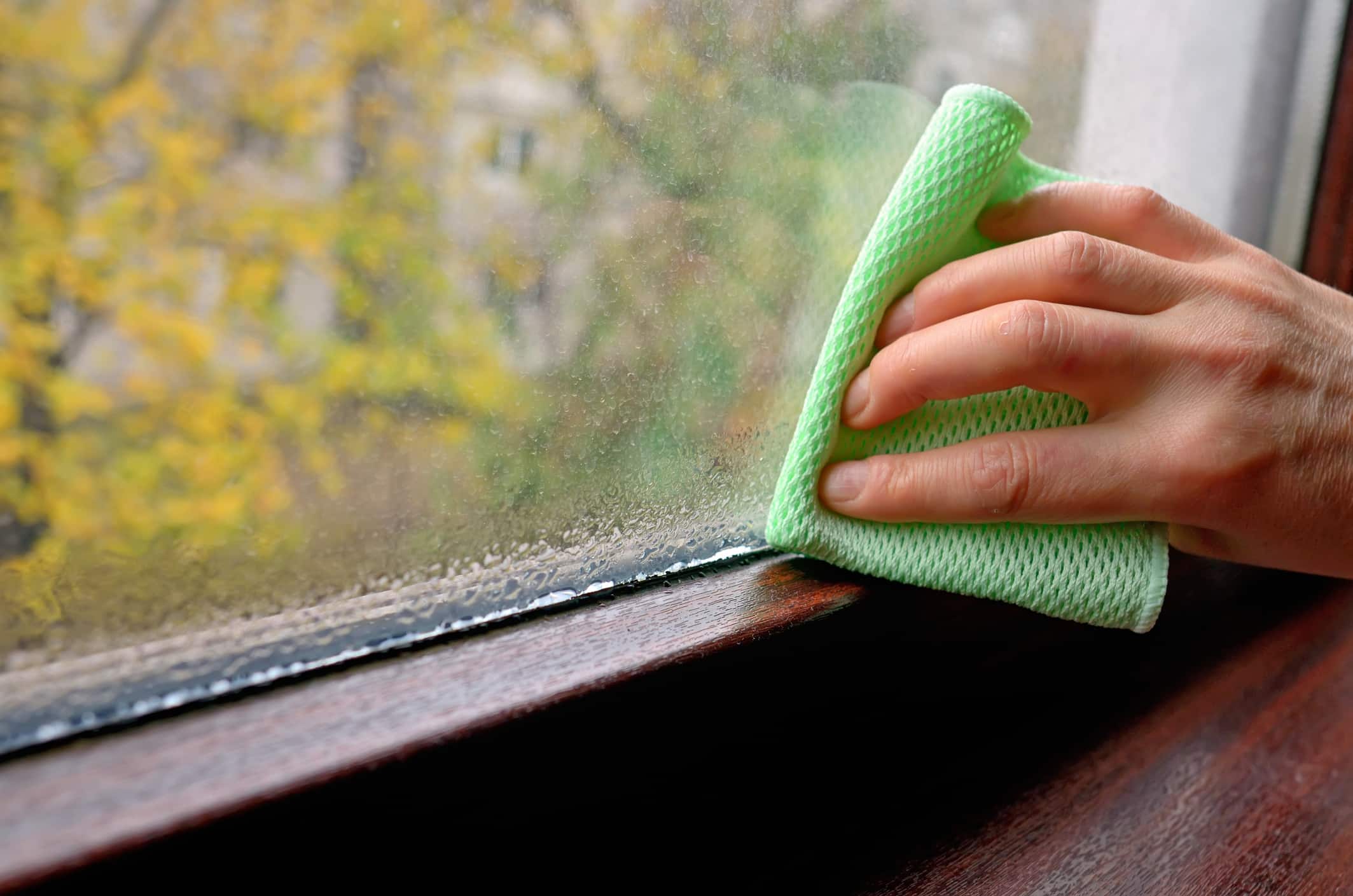 Homeowner wiping condensation from a window interior, a common sign of high indoor humidity in summer.