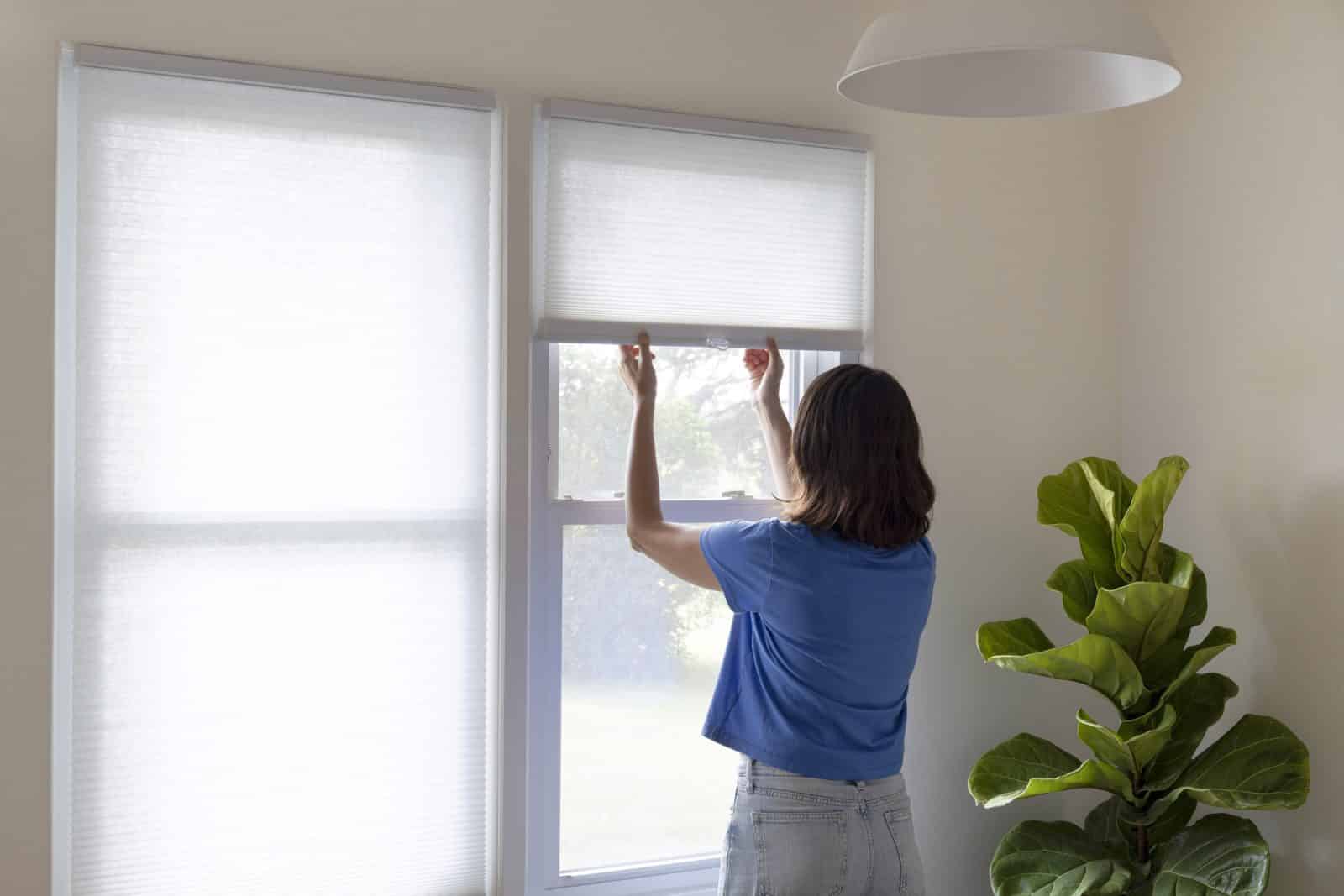A woman lowering window shades to block summer sun and reduce heat gain in her home.